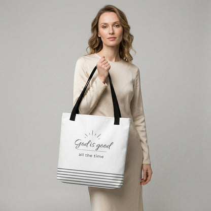 Model holding a white tote bag with black handles that reads “God is good all the time” in dark gray with a striped bottom design.