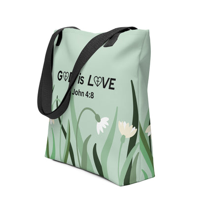 Angled view of sage green tote bag featuring “GOD is LOVE” and “1 John 4:8” over a floral-and-grass illustration, with black straps.