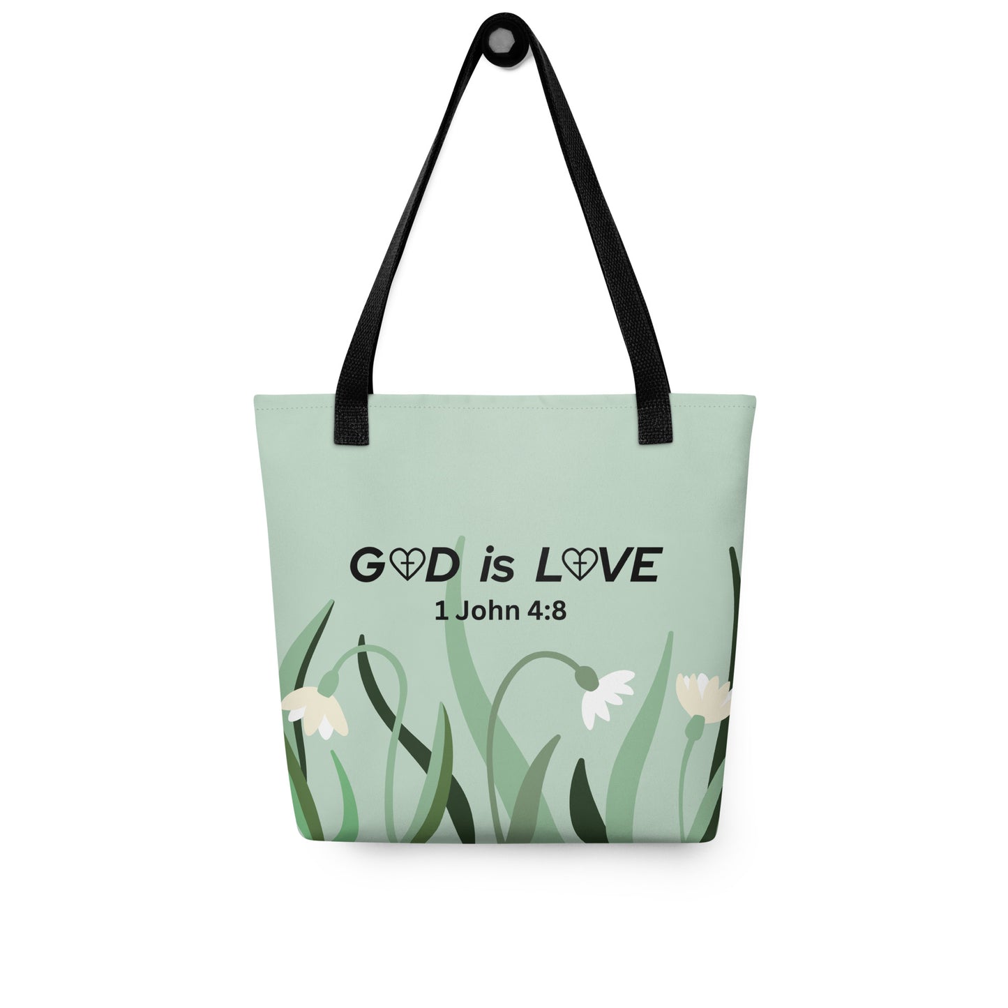 Sage green tote bag hanging on a hook, printed with “GOD is LOVE” and “1 John 4:8” above a simple flower and grass design, with black straps.
