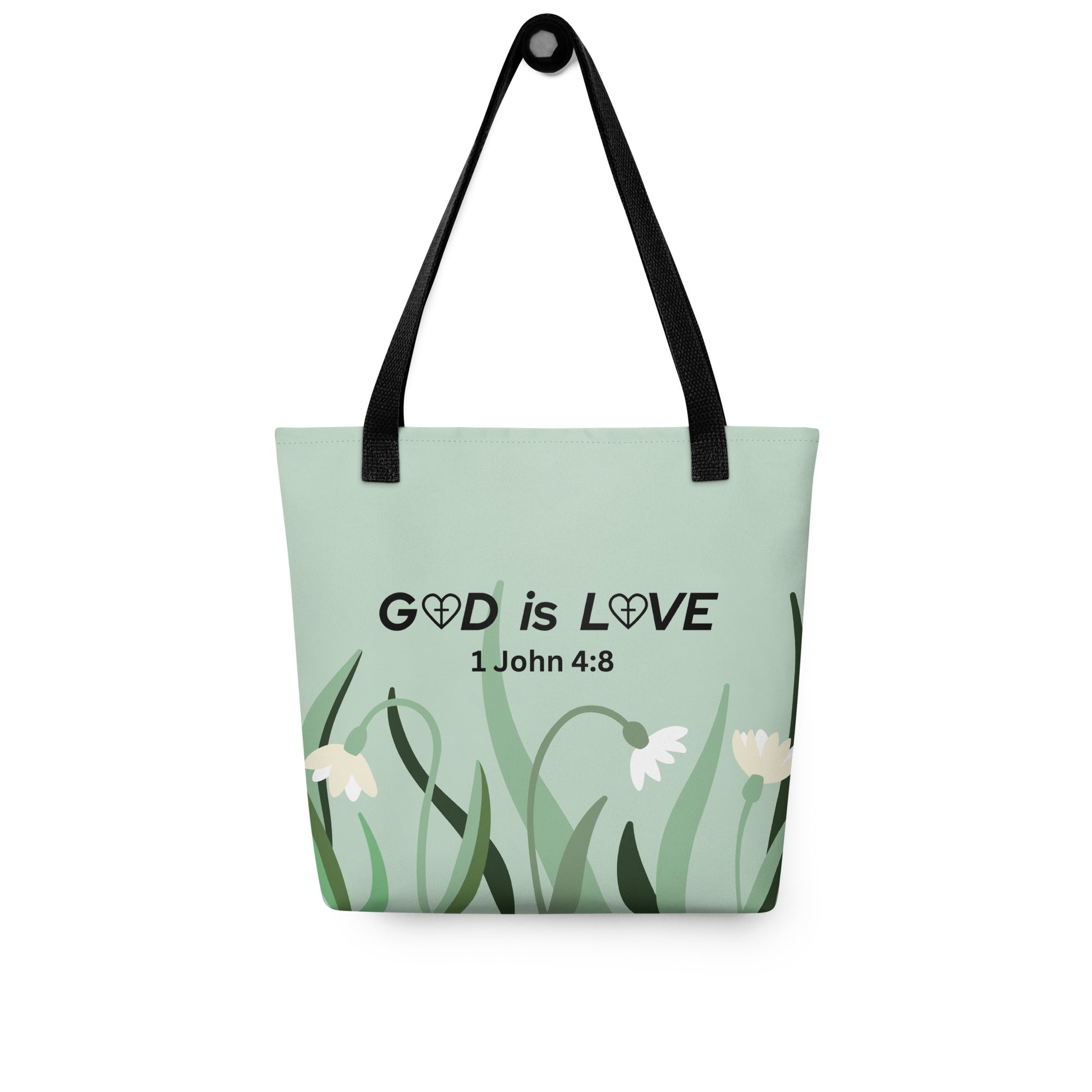 Sage green tote bag hanging on a hook, printed with “GOD is LOVE” and “1 John 4:8” above a simple flower and grass design, with black straps.