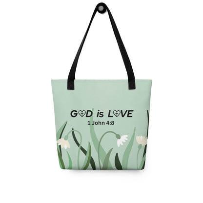 Sage green tote bag hanging on a hook, printed with “GOD is LOVE” and “1 John 4:8” above a simple flower and grass design, with black straps.