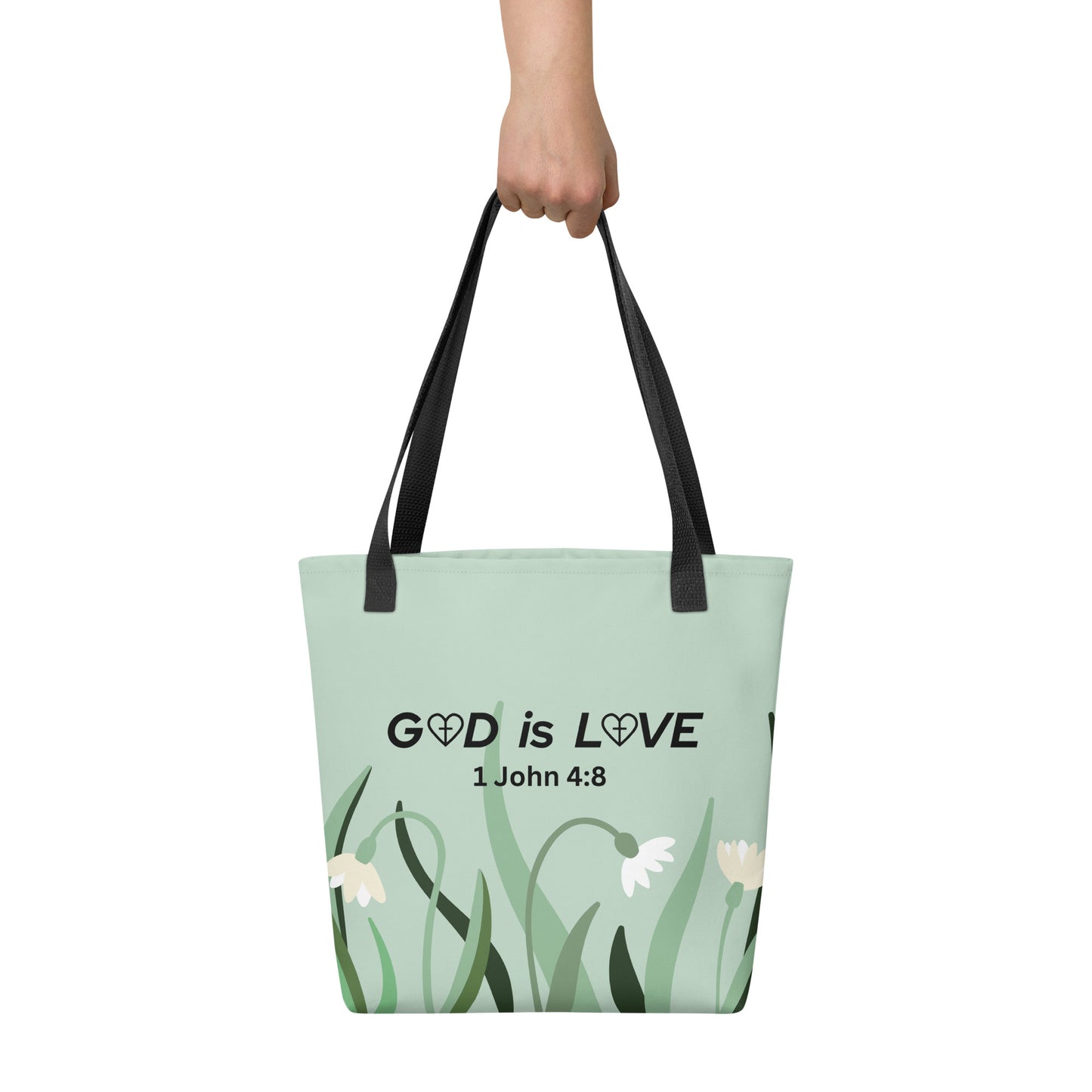 Hand holding sage green tote bag with “GOD is LOVE” and “1 John 4:8” Bible verse design and minimalist flowers, with black straps.