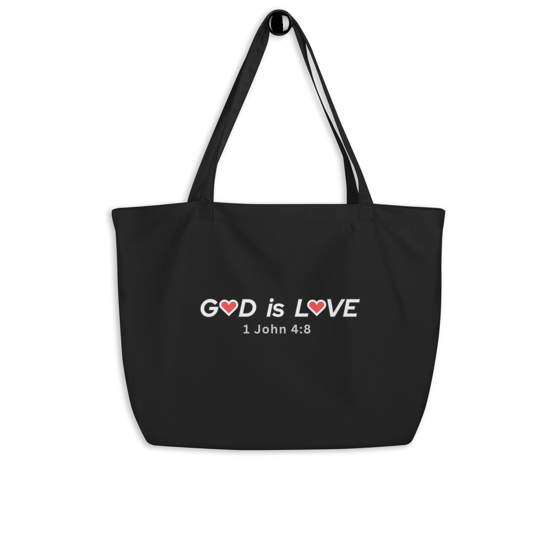 Large black cotton tote bag hanging on a hook featuring “GOD is LOVE” with red heart accents and “1 John 4:8” below.
