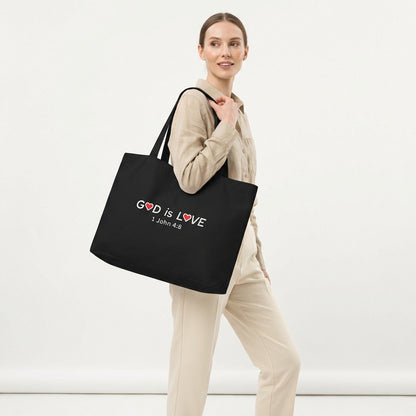 Model holding a large black cotton tote bag printed with “GOD is LOVE,” red heart accents, and “1 John 4:8.”