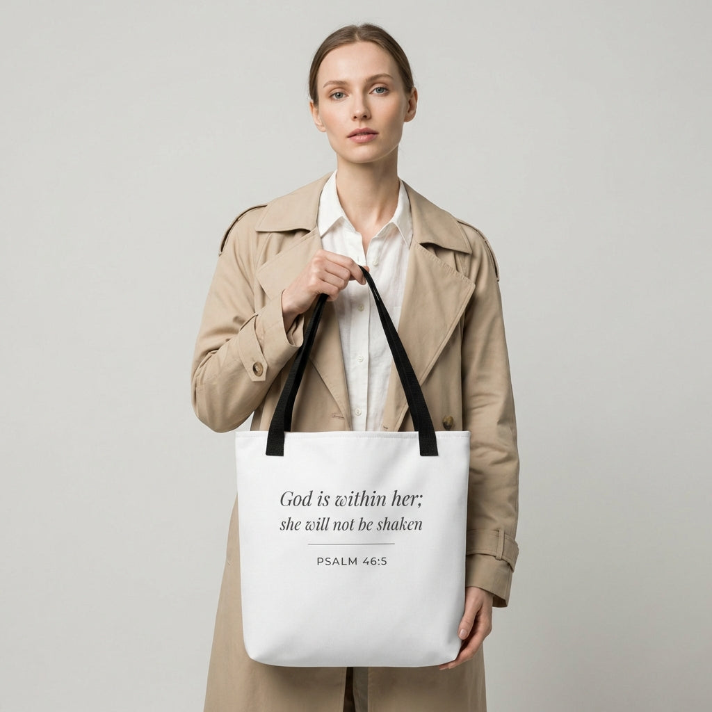 Lifestyle photo of a model holding a white tote bag featuring ‘God is within her; she will not be shaken’ (Psalm 46:5) in dark gray.