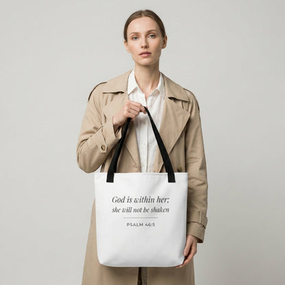 Lifestyle photo of a model holding a white tote bag featuring ‘God is within her; she will not be shaken’ (Psalm 46:5) in dark gray.