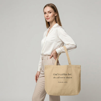 Model carrying a natural cotton Christian tote bag with beige handles and “God is within her; she will not be shaken” scripture text.
