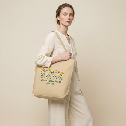 Model carrying a beige cotton tote bag with floral artwork and the text “Grace Upon Grace” and “John 1:16.”