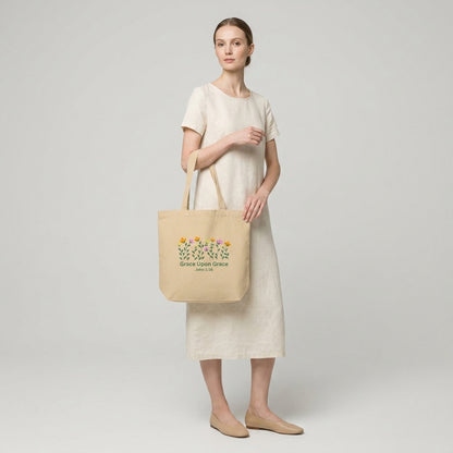 Model carrying a beige cotton tote bag with floral artwork and the text “Grace Upon Grace” and “John 1:16.”