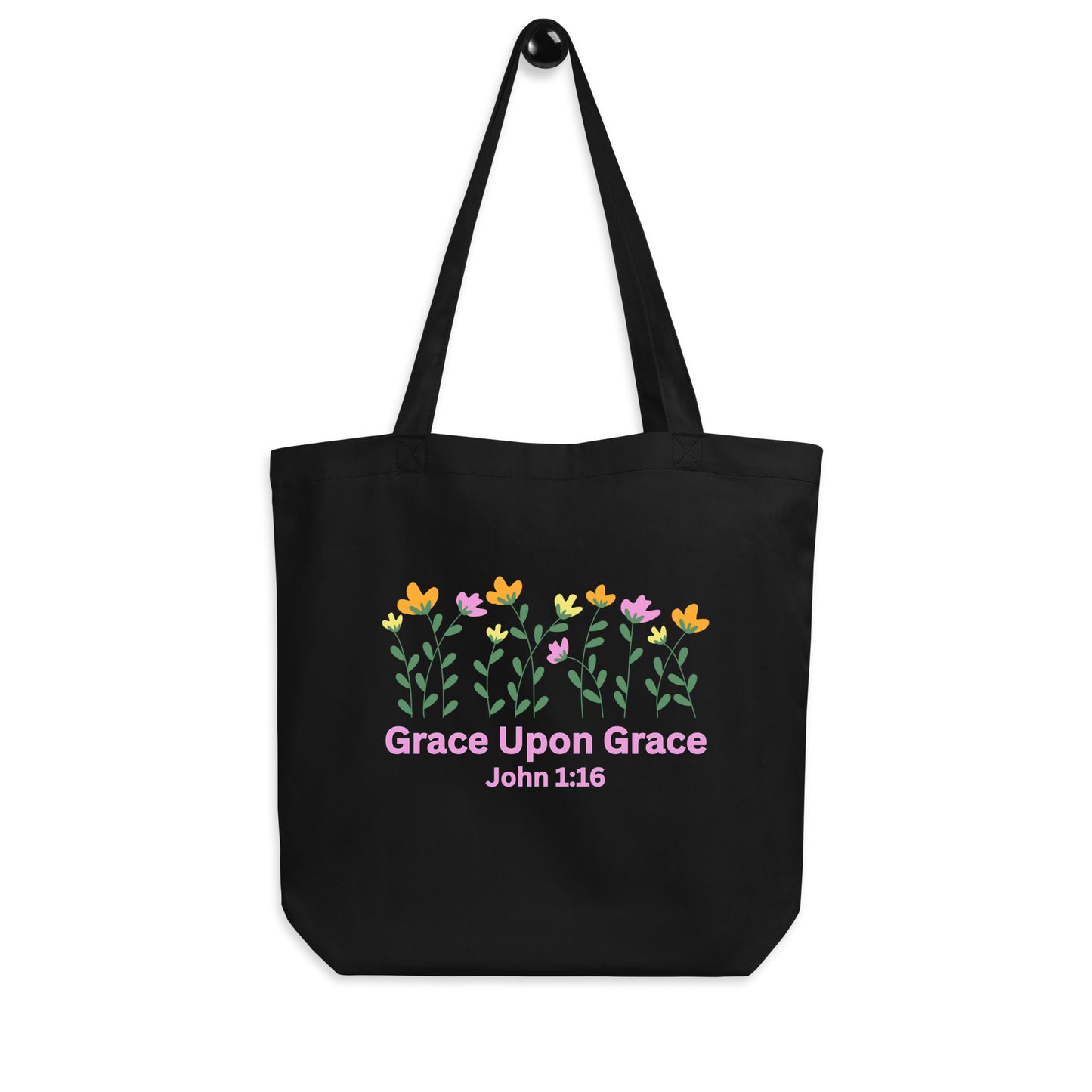 Black cotton tote bag hanging on a hook featuring the floral design and the pink “Grace Upon Grace” John 1:16 print.
