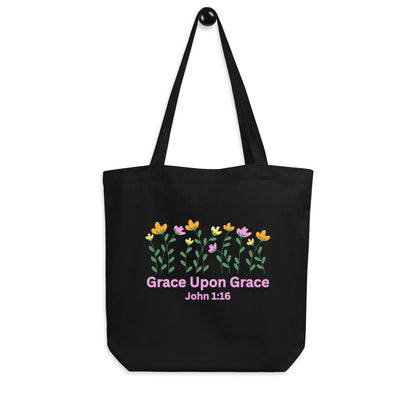 Black cotton tote bag hanging on a hook featuring the floral design and the pink “Grace Upon Grace” John 1:16 print.