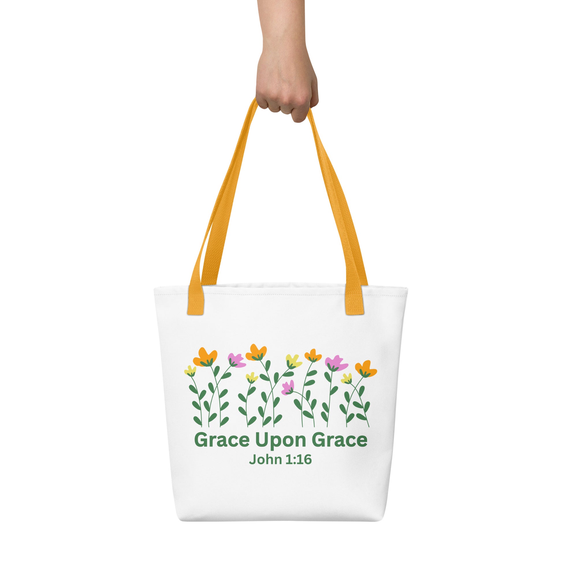 A hand holding the white ‘Grace Upon Grace’ John 1:16 tote bag with mustard-yellow straps; floral graphic and green text visible.