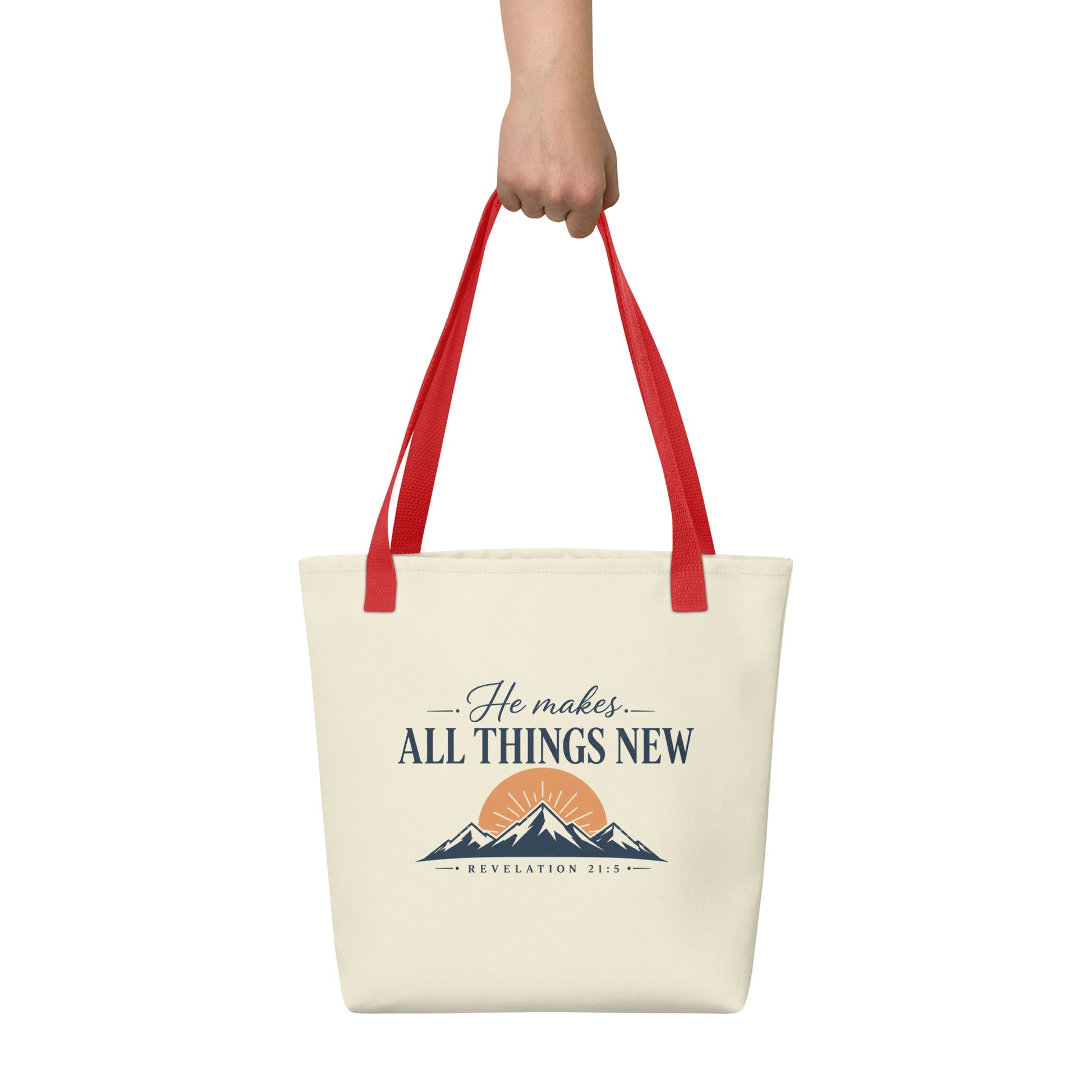 Person holding a cream Christian tote bag with red handles, a mountain sunrise design, and the Bible verse reference Revelation 21:5.