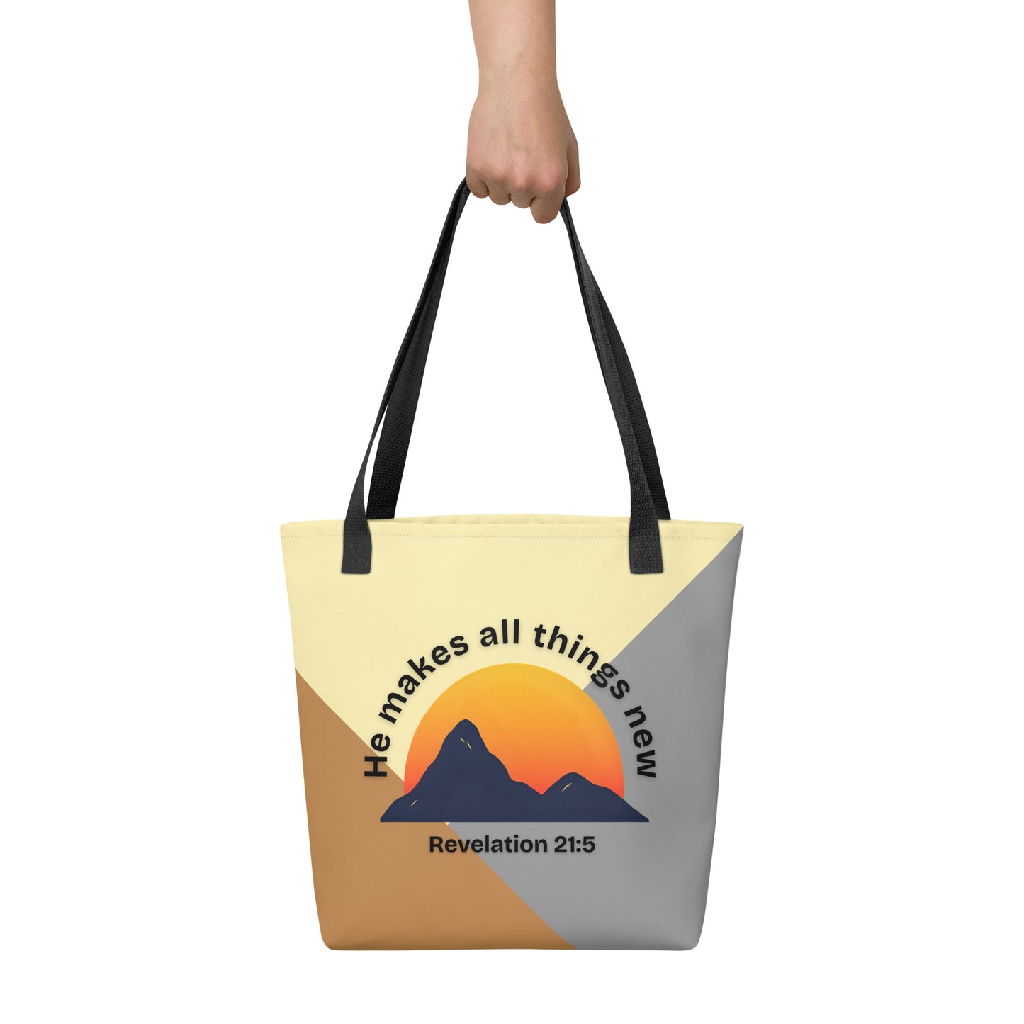 Tote bag held by hand with black straps, featuring neutral color-block panels and a mountain sunset graphic with the words “He makes all things new” and “Revelation 21:5.”