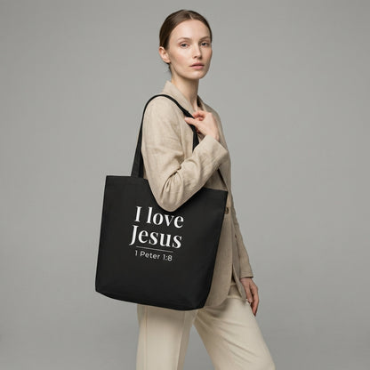 Model holding a black cotton tote bag printed with “I love Jesus” and “1 Peter 1:8” in white text.