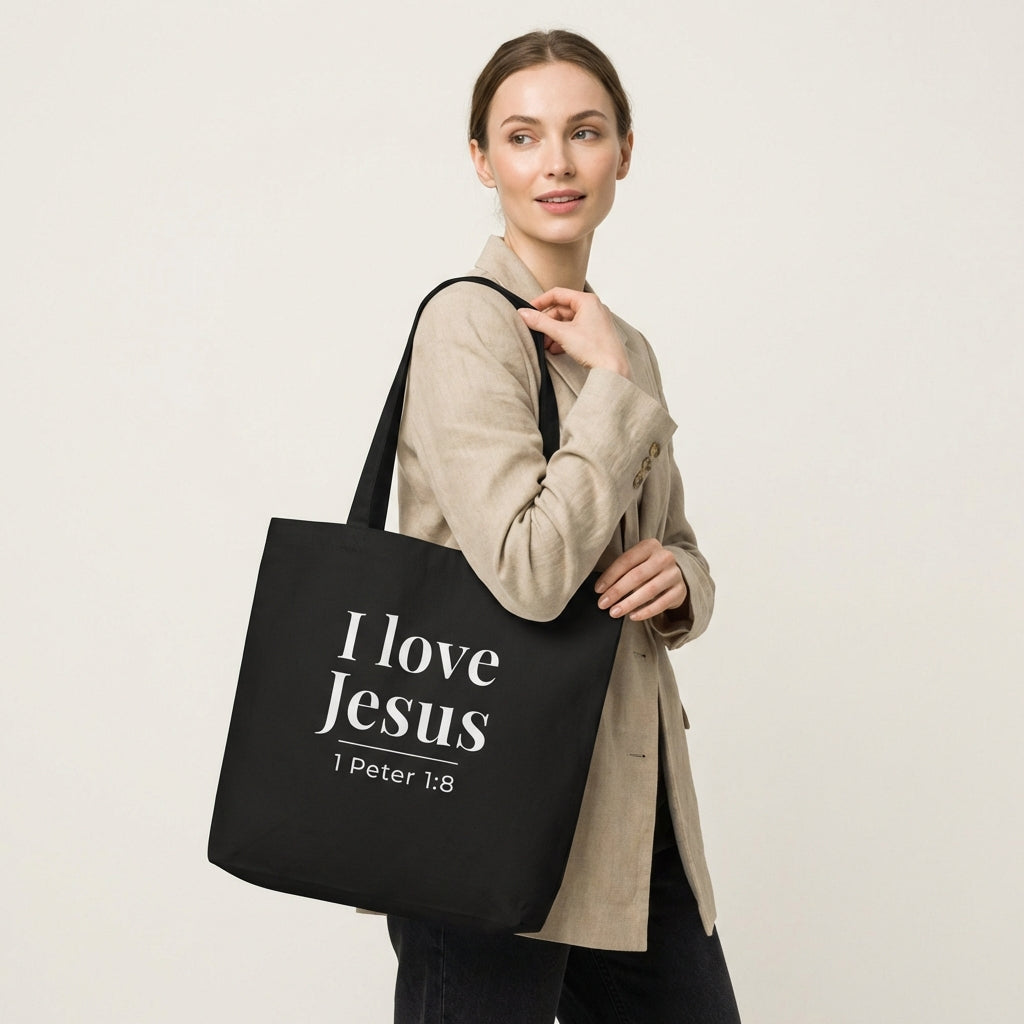 Model holding a black cotton tote bag printed with “I love Jesus” and “1 Peter 1:8” in white text.