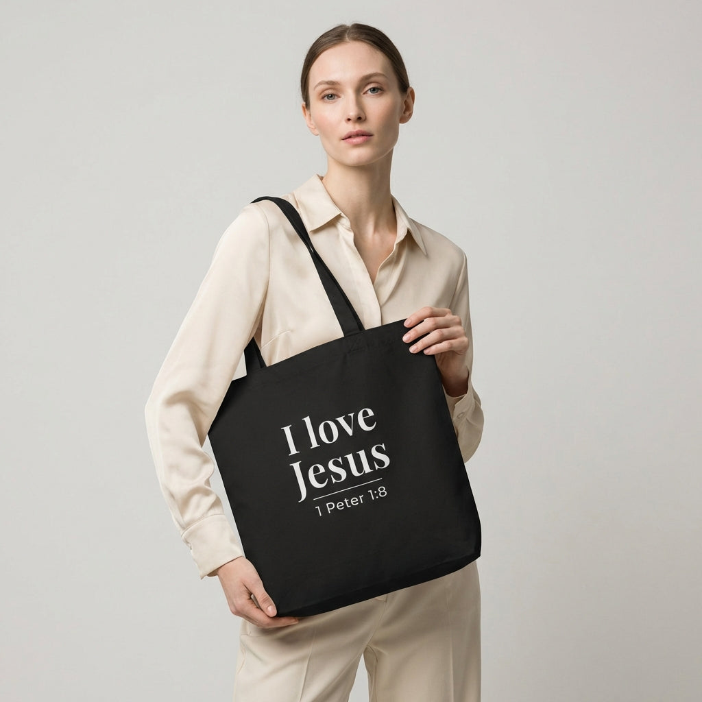 Model holding a black cotton tote bag printed with “I love Jesus” and “1 Peter 1:8” in white text.