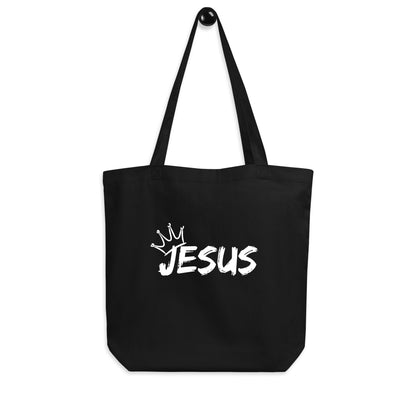Black cotton tote bag hanging on a hook featuring a white “JESUS” design with a crown graphic above it.