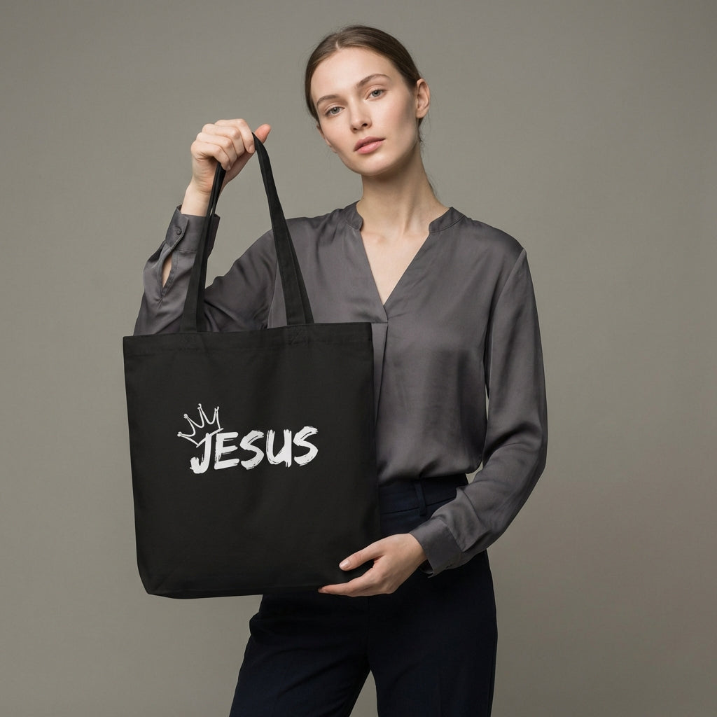 Model holding a black cotton tote bag with a white “JESUS” graphic and crown detail on the front.