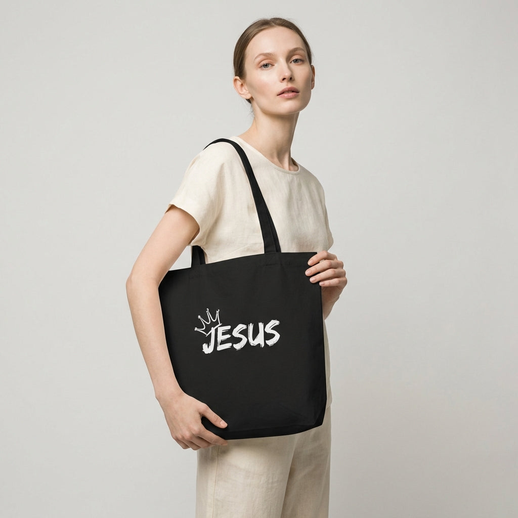 Model holding a black cotton tote bag with a white “JESUS” graphic and crown detail on the front.