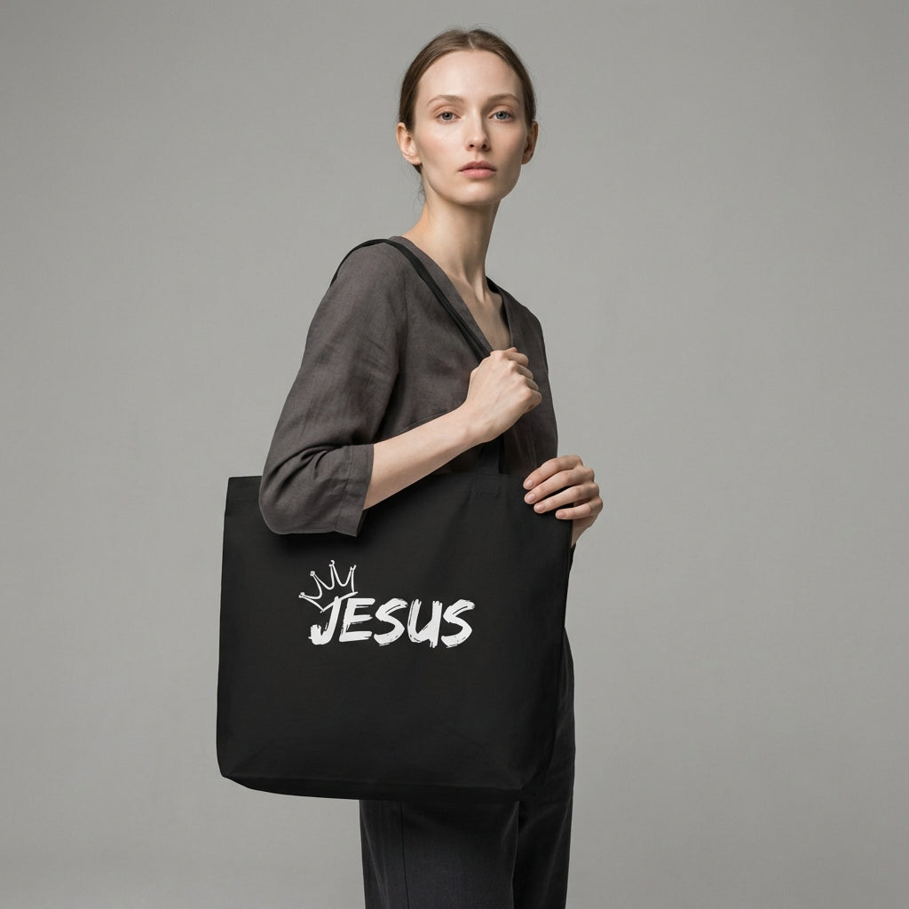 Model holding a black cotton tote bag with a white “JESUS” graphic and crown detail on the front.