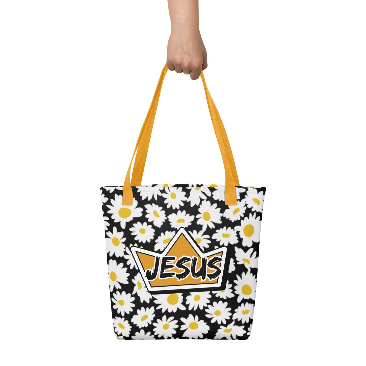 Tote bag held by hand with mustard yellow straps, featuring a black background with white daisies and a crown emblem with “JESUS” across the center.