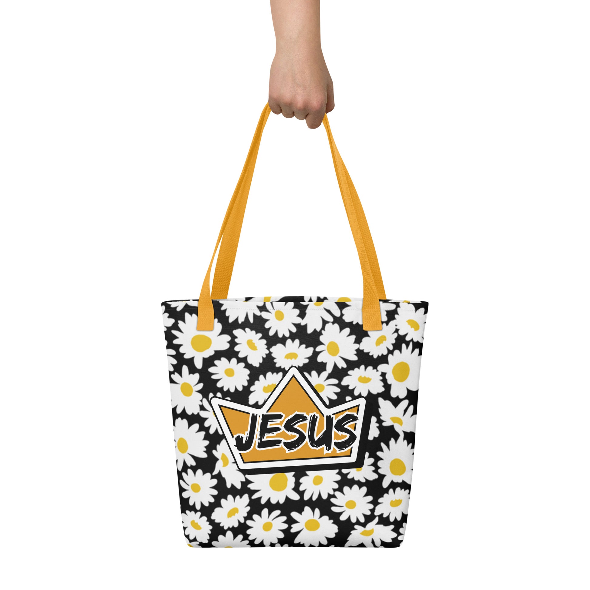 Tote bag held by hand with mustard yellow straps, featuring a black background with white daisies and a crown emblem with “JESUS” across the center.