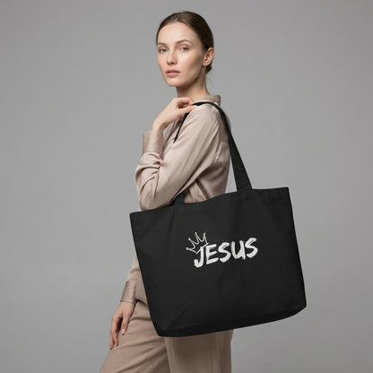 Model holding a large black cotton tote bag with a white “JESUS” graphic and crown detail on the front.