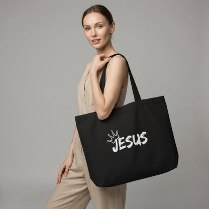 Model holding a large black cotton tote bag with a white “JESUS” graphic and crown detail on the front.