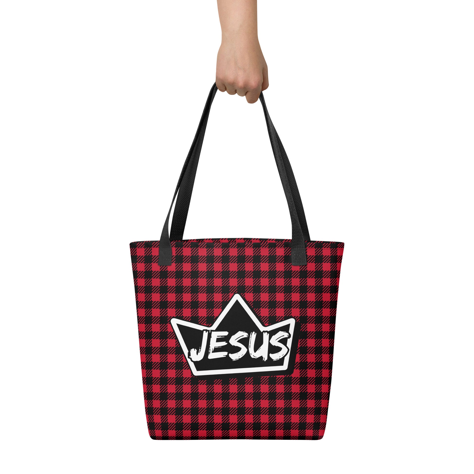 Tote bag held by hand with black straps, featuring a red and black buffalo check pattern and a bold crown emblem with “JESUS” across the center.