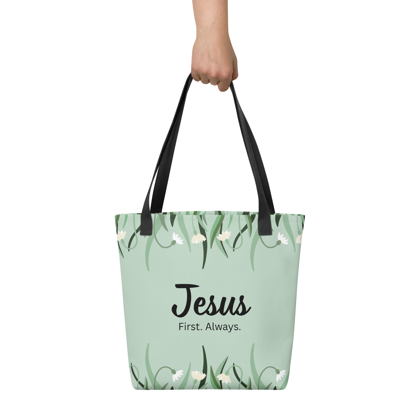 Tote bag held by hand with black straps, featuring a mint green floral pattern and the words “Jesus. First. Always.”