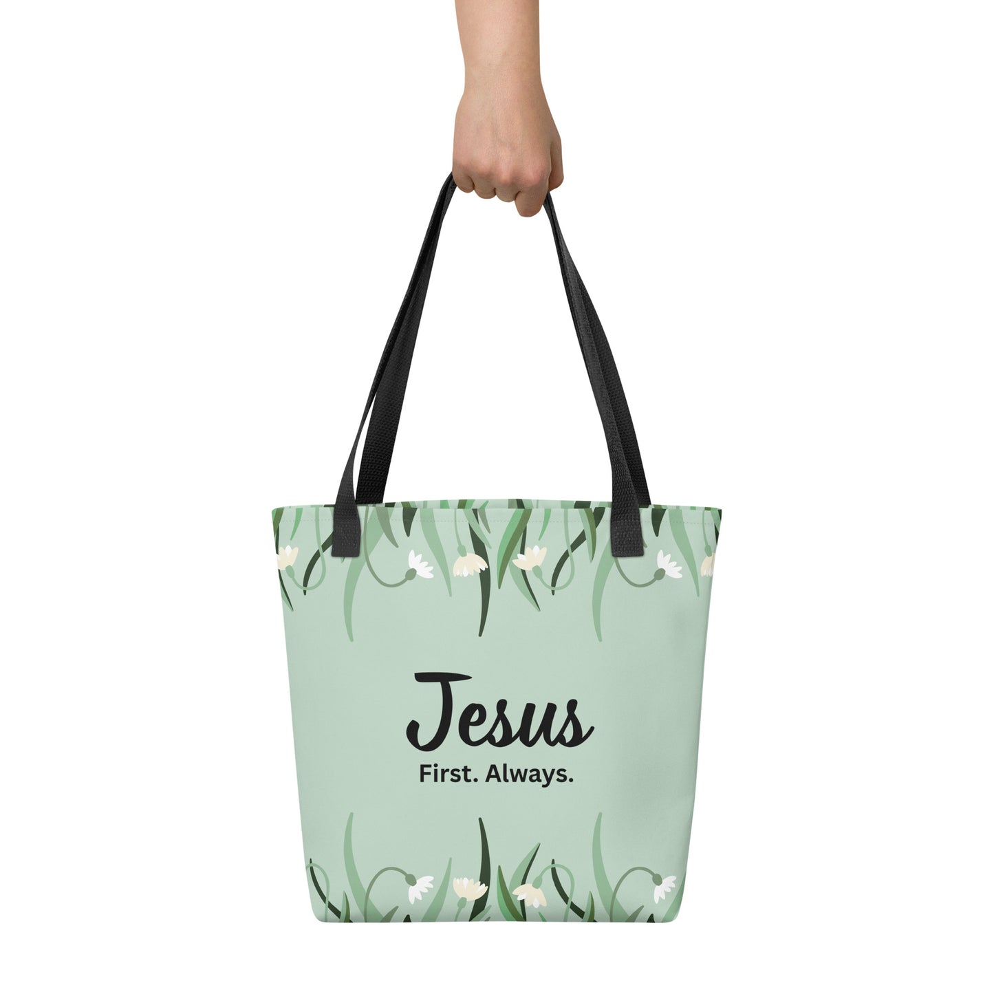 Tote bag held by hand with black straps, featuring a mint green floral pattern and the words “Jesus. First. Always.”