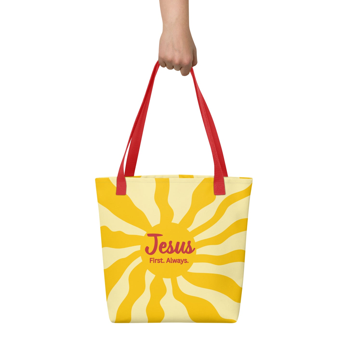 Hand holding red-handle tote bag with yellow sunburst design and “Jesus First. Always.” message.