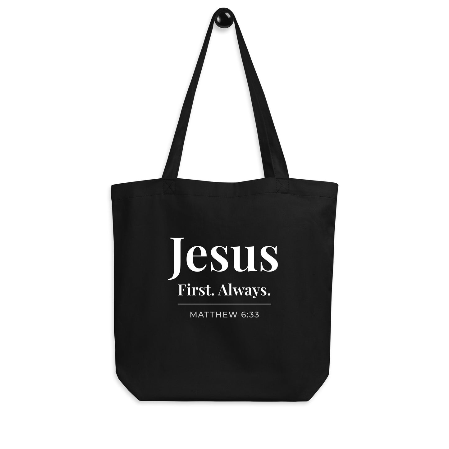 Black cotton tote bag hanging on a hook featuring “Jesus First. Always.” and “Matthew 6:33” in white text.