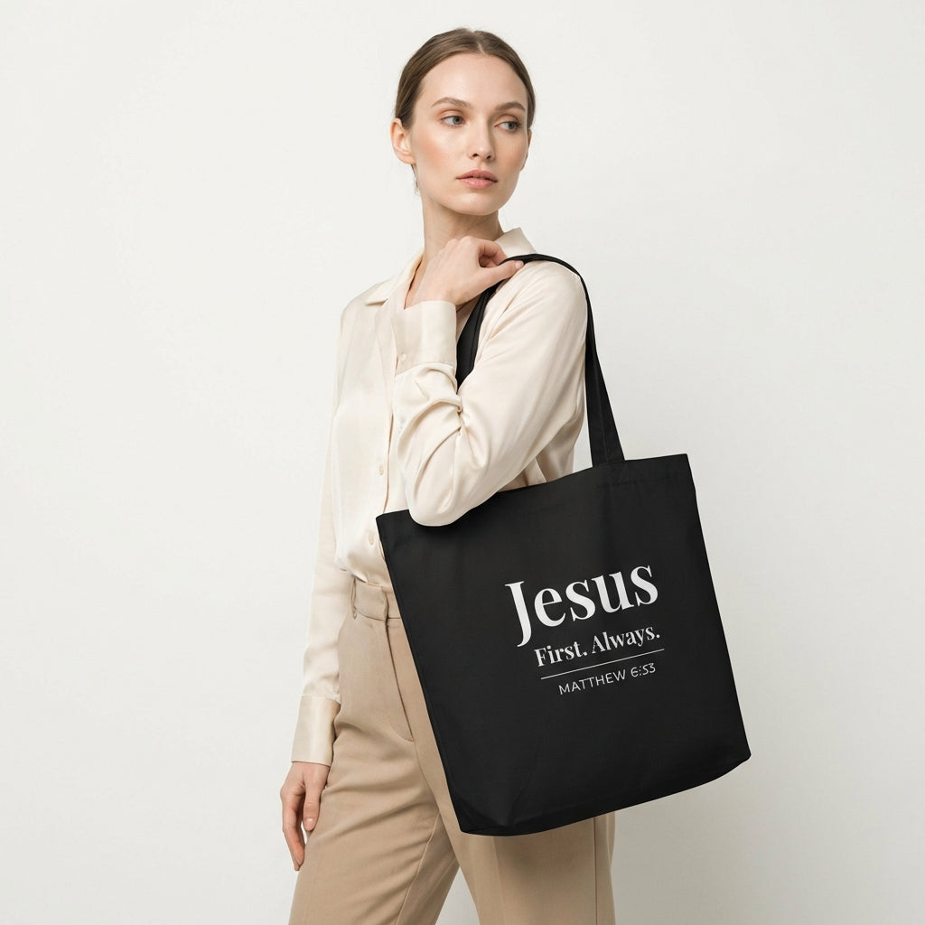 Model holding a black cotton tote bag printed with “Jesus First. Always.” and “Matthew 6:33” in white text.