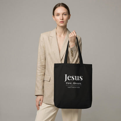 Model holding a black cotton tote bag printed with “Jesus First. Always.” and “Matthew 6:33” in white text.
