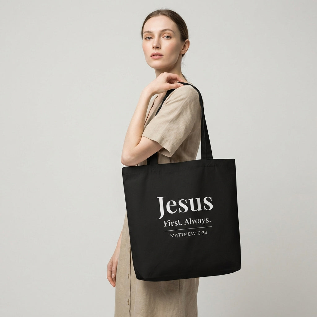 Model holding a black cotton tote bag printed with “Jesus First. Always.” and “Matthew 6:33” in white text.