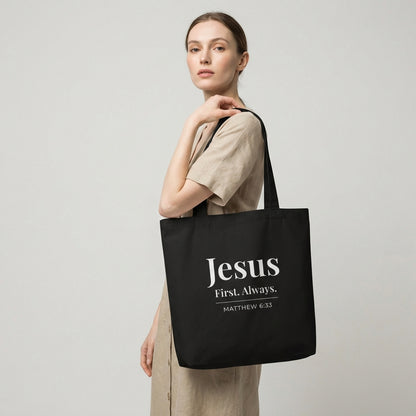 Model holding a black cotton tote bag printed with “Jesus First. Always.” and “Matthew 6:33” in white text.