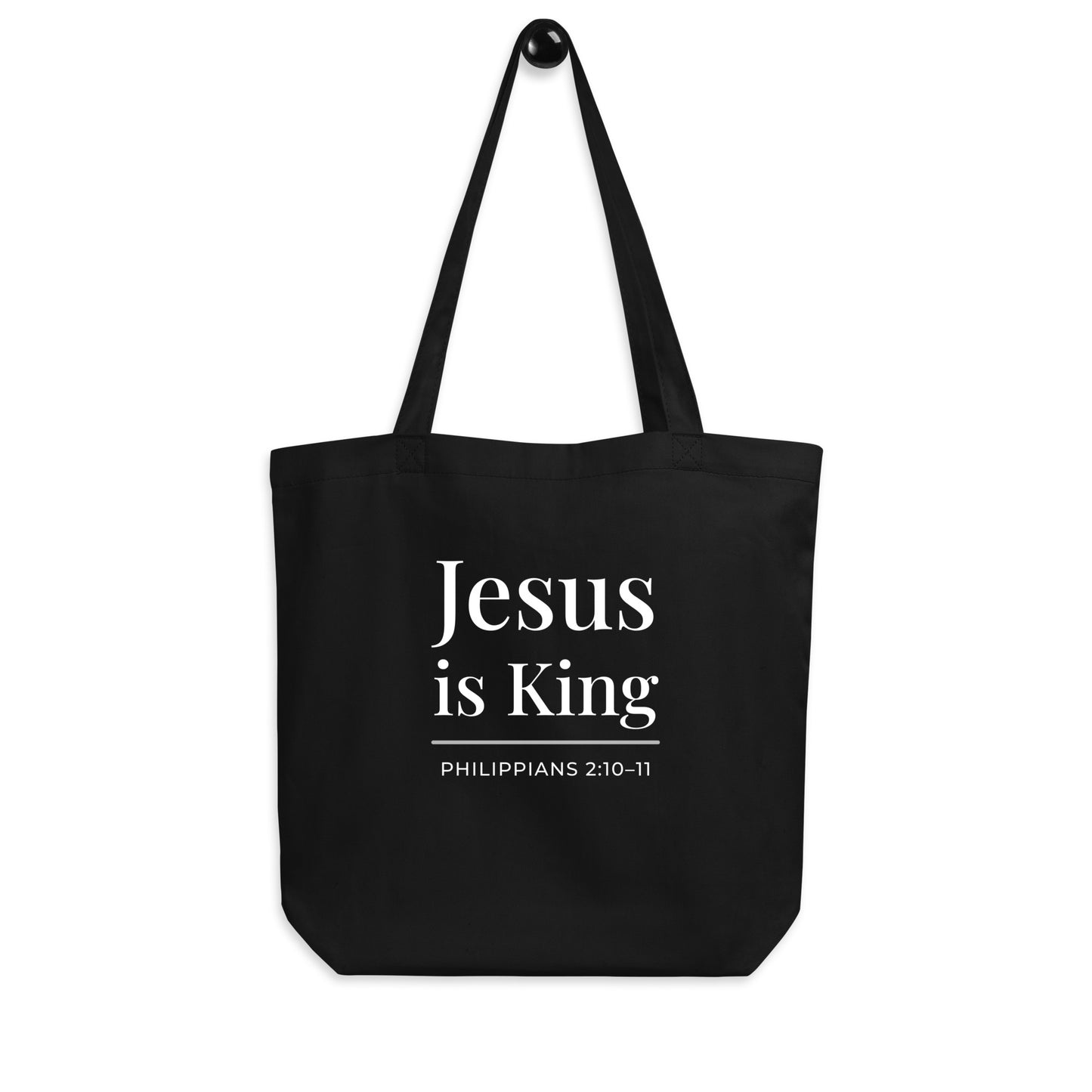 Black tote bag hanging on a hook featuring white “Jesus is King” text and “Philippians 2:10–11” below.