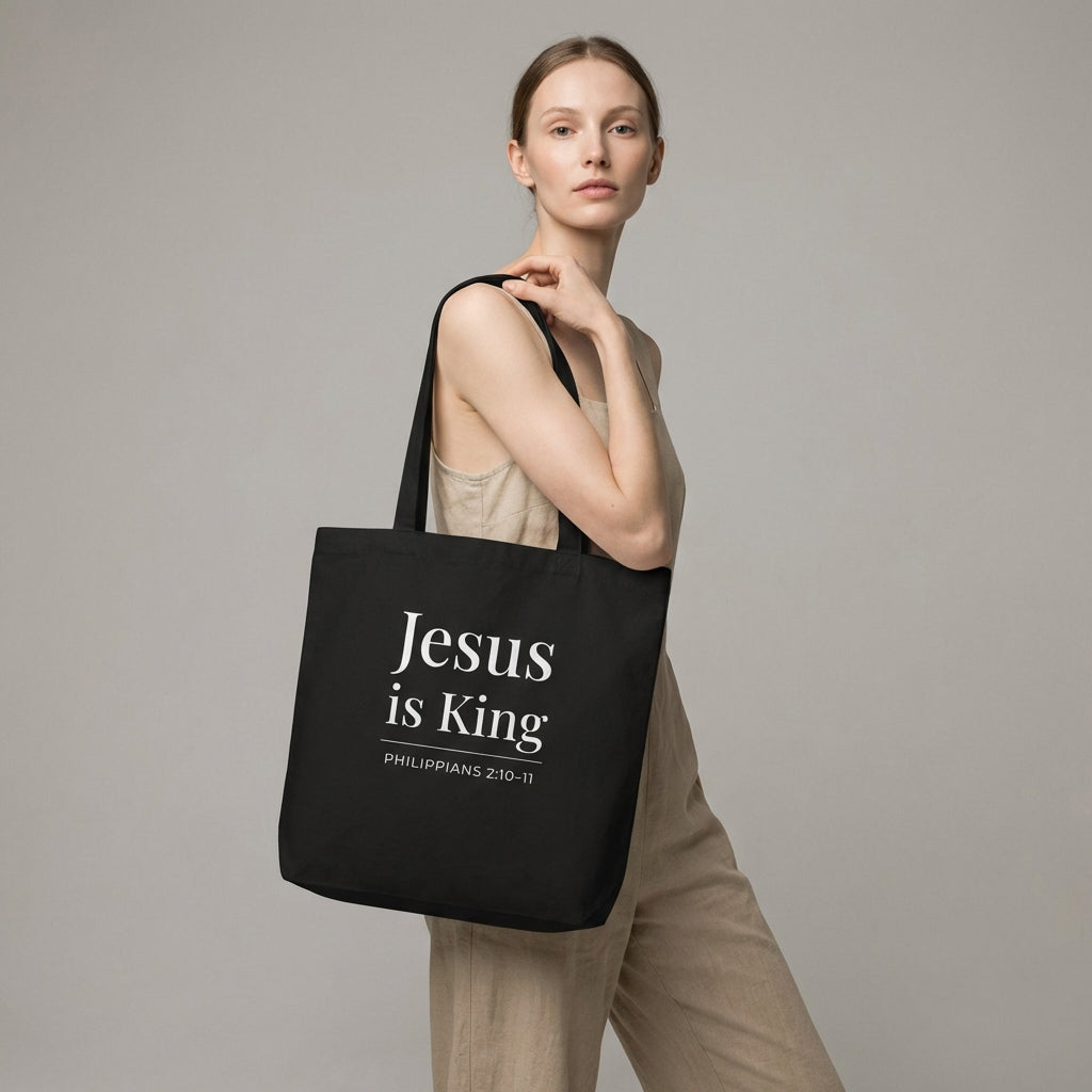 Model holding a black tote bag with white “Jesus is King” text and “Philippians 2:10–11” design.