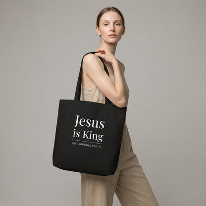 Model holding a black tote bag with white “Jesus is King” text and “Philippians 2:10–11” design.