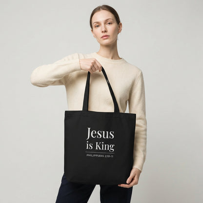Model holding a black tote bag with white “Jesus is King” text and “Philippians 2:10–11” design.