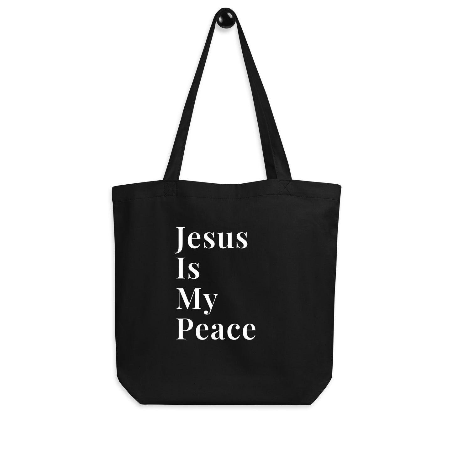 Black cotton tote bag hanging on a hook with white “Jesus Is My Peace” text on the front.