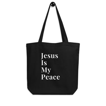 Black cotton tote bag hanging on a hook with white “Jesus Is My Peace” text on the front.