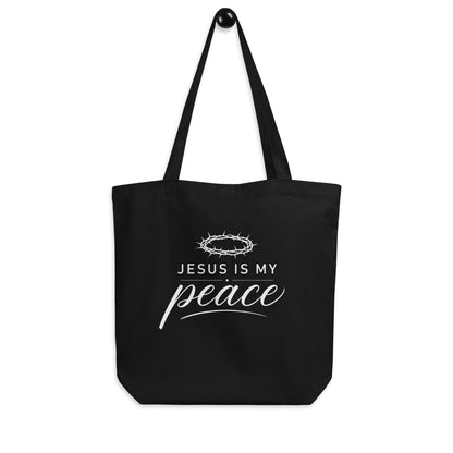 Black Christian cotton tote bag hanging on a hook, featuring matching black straps and a white “Jesus Is My Peace” design.