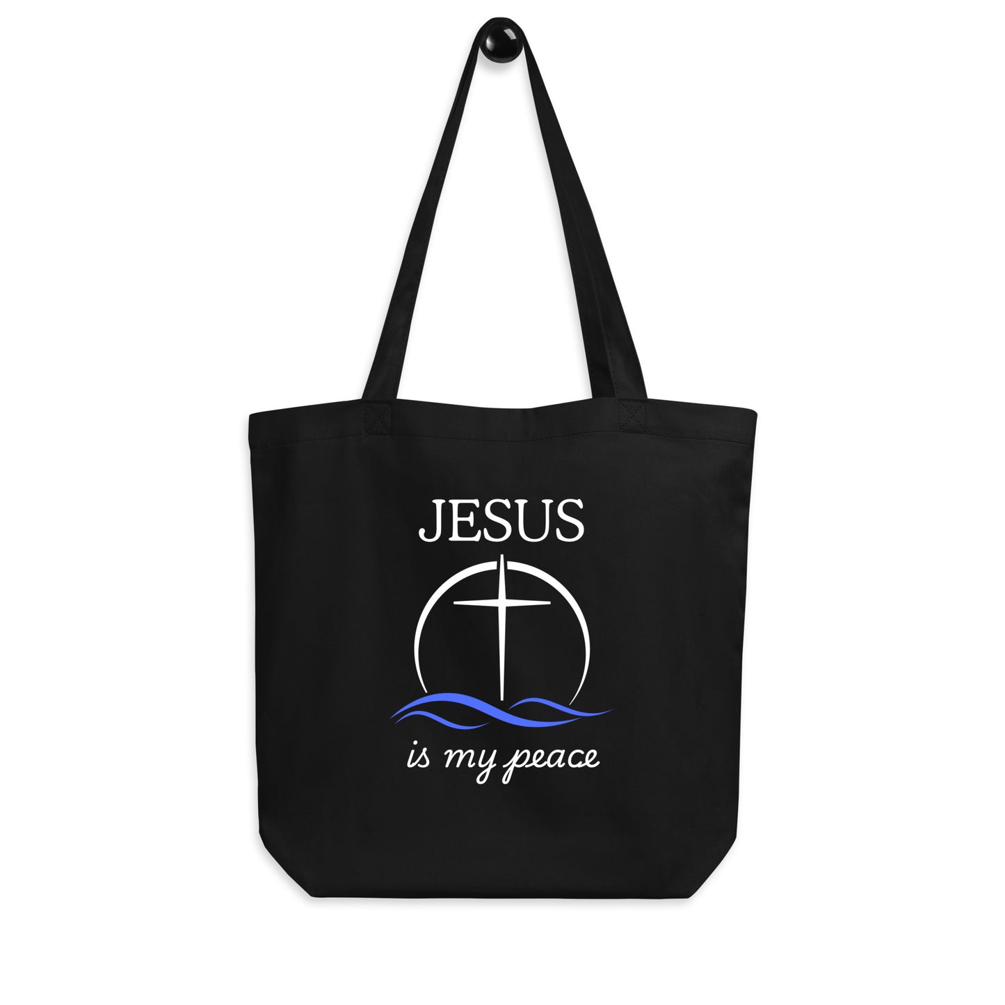 Black cotton tote bag hanging on a hook featuring “JESUS,” a cross-in-circle design with blue waves, and “is my peace” in white lettering.