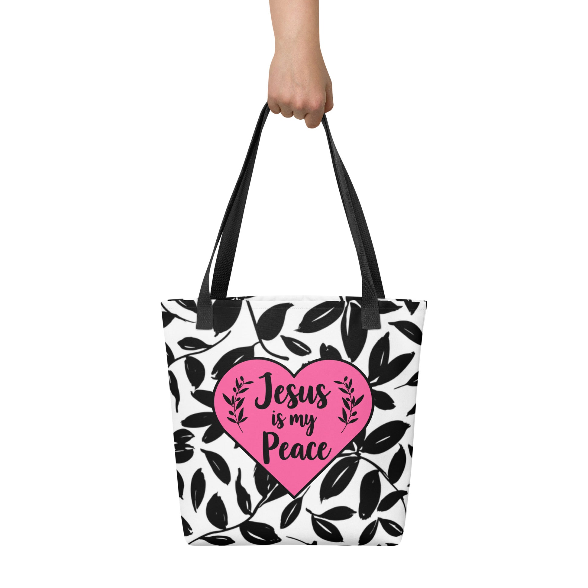 Handheld view of tote bag with black leaf pattern, black straps, and pink heart centerpiece that says “Jesus is my Peace.”