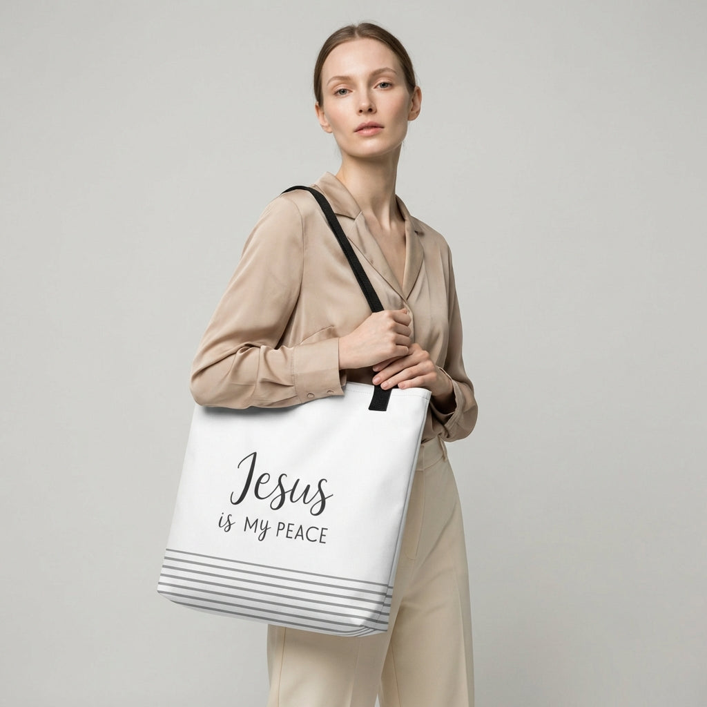 Model holding a white tote bag with black handles that reads “Jesus is my peace” in dark gray with a striped bottom design.