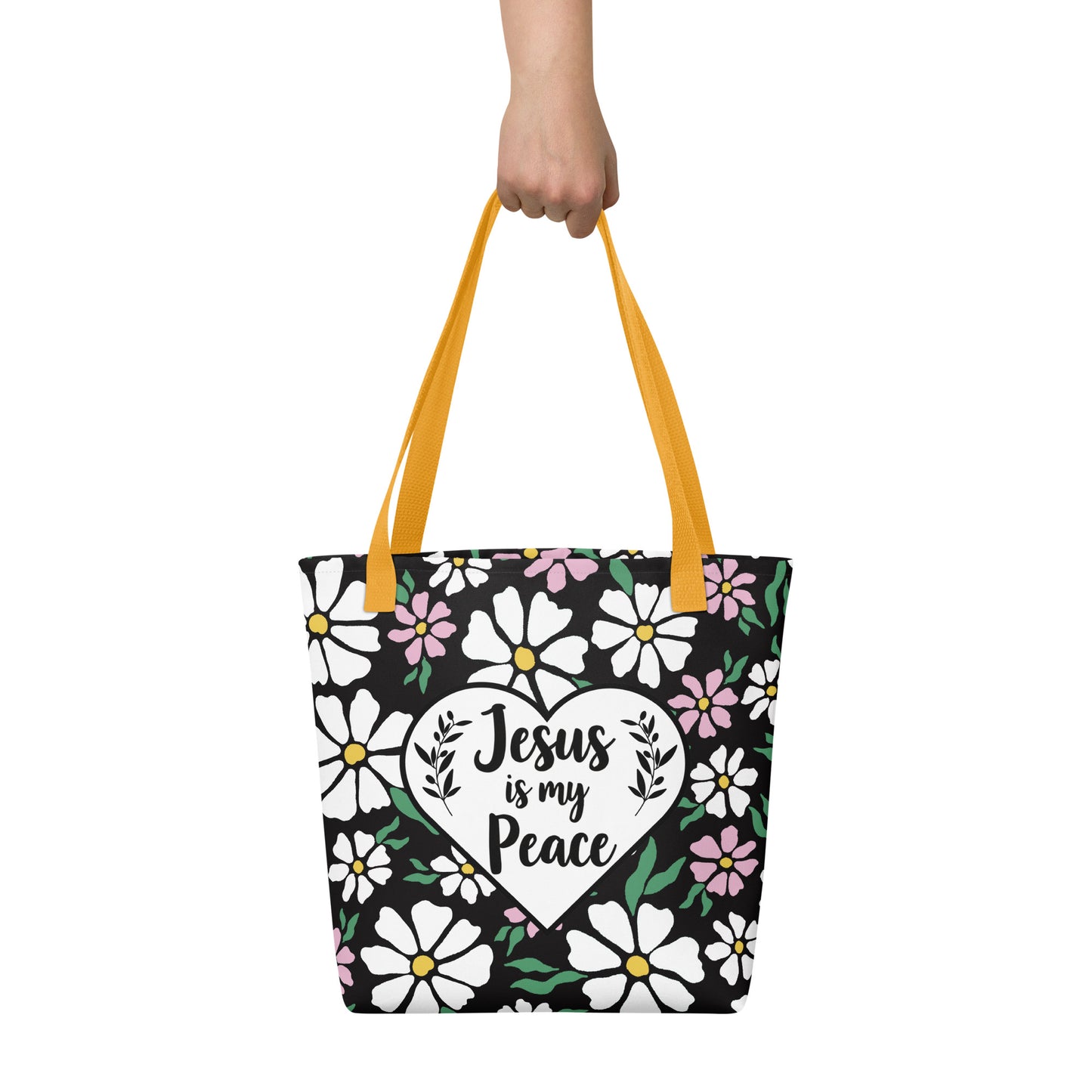 Tote bag held by hand with mustard yellow straps, featuring a black background with white flowers and a heart-shaped “Jesus is my Peace” message.