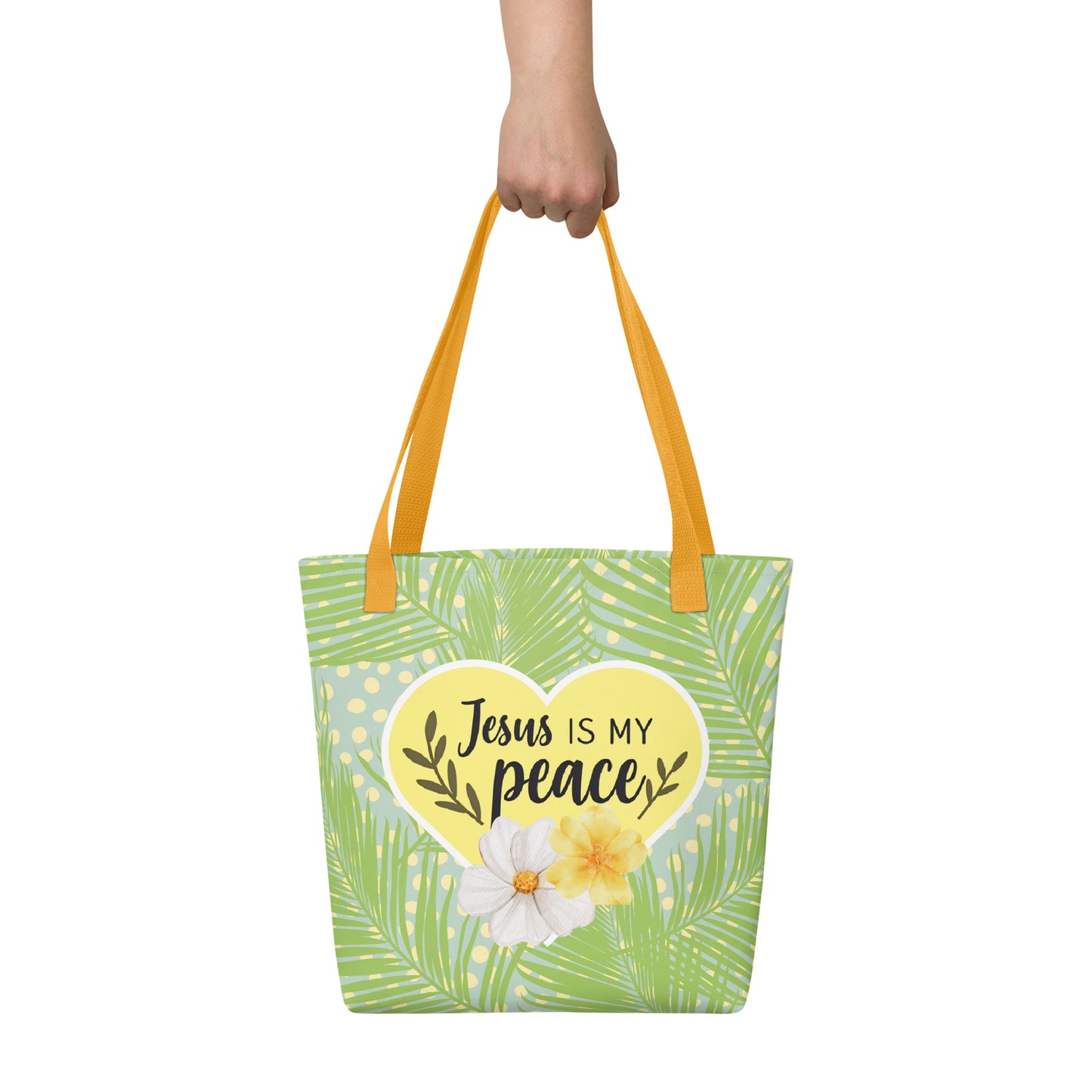 Handheld view of the tote bag with yellow handles and tropical palm-leaf pattern, featuring a yellow heart that reads “Jesus is my peace” with flowers.